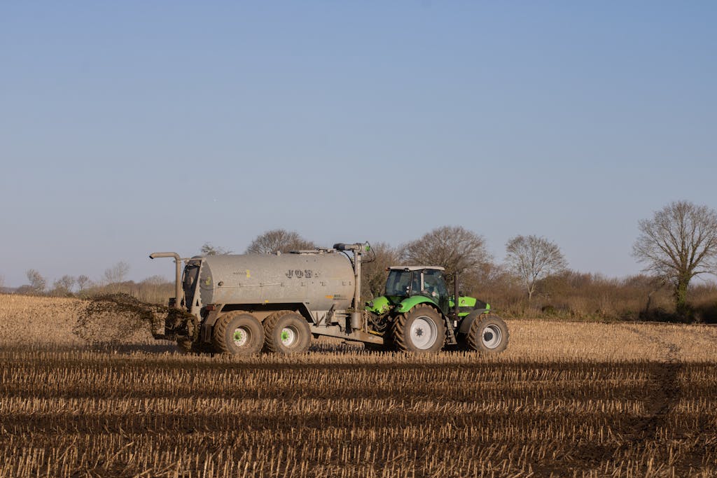 A green tractor spreading fertilizer on a brown, freshly plowed farmland under a clear sky.